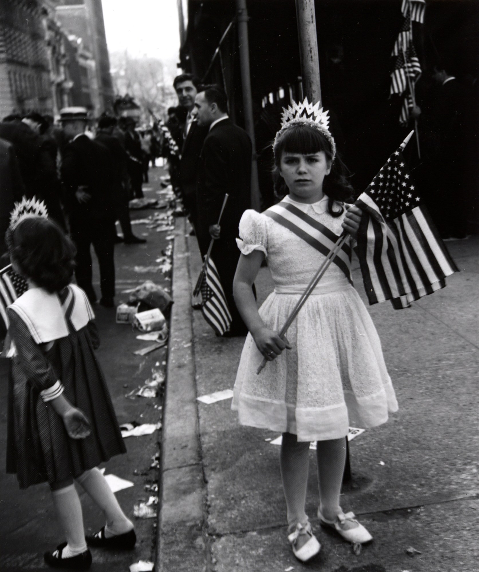 a girl in a dainty dress and ballet shoes on the street wears a crown and holds an american flag with two hands. she has a crown on.