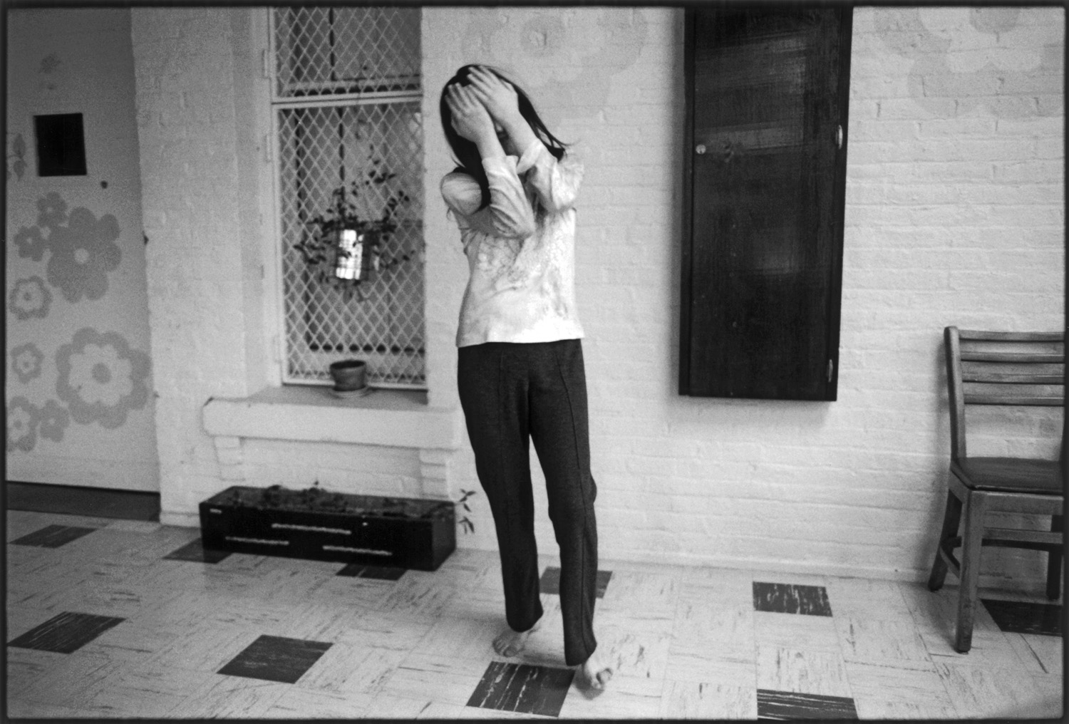 A woman standing barefoot on checkered tile, holding her hands up to her face