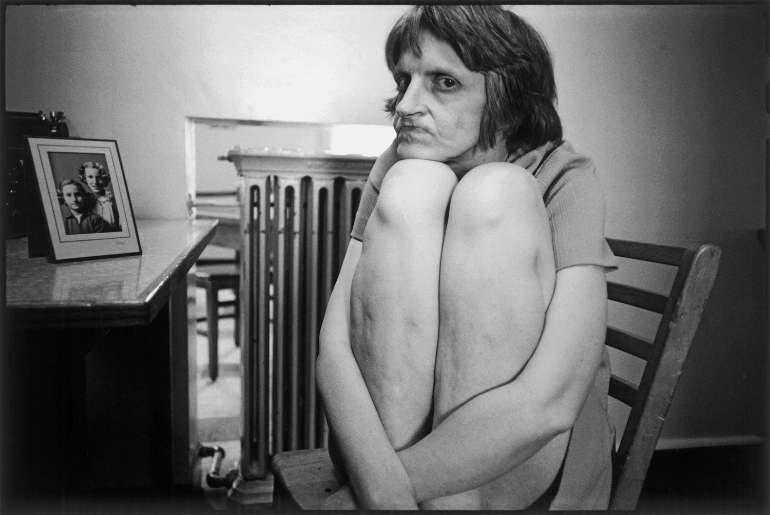 I woman sitting in a wooden chair next to a framed photograph holding her knees