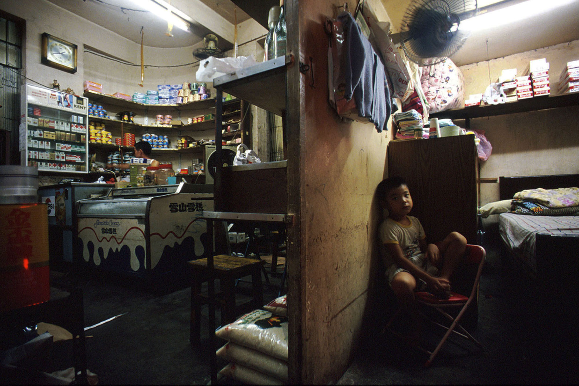 A child sits on a chair while on the other side of the wall a man works in a shop within Kowloon Walled City