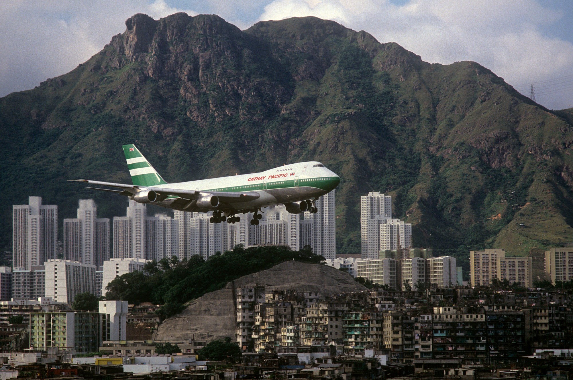 A plane flies low over Kowloon Walled City with mountains in the background