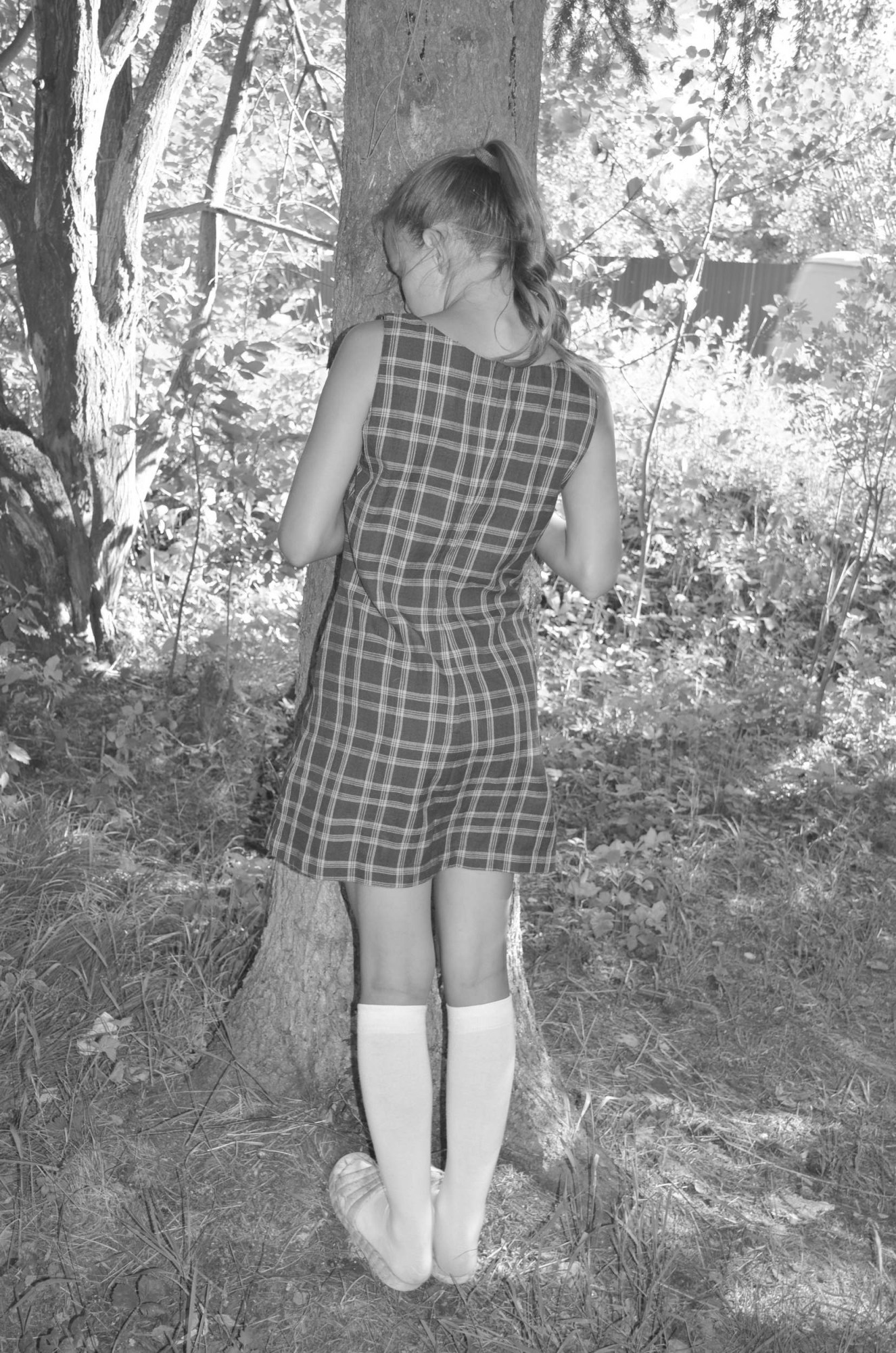 a girl in a checked dress with school socks faces a tree