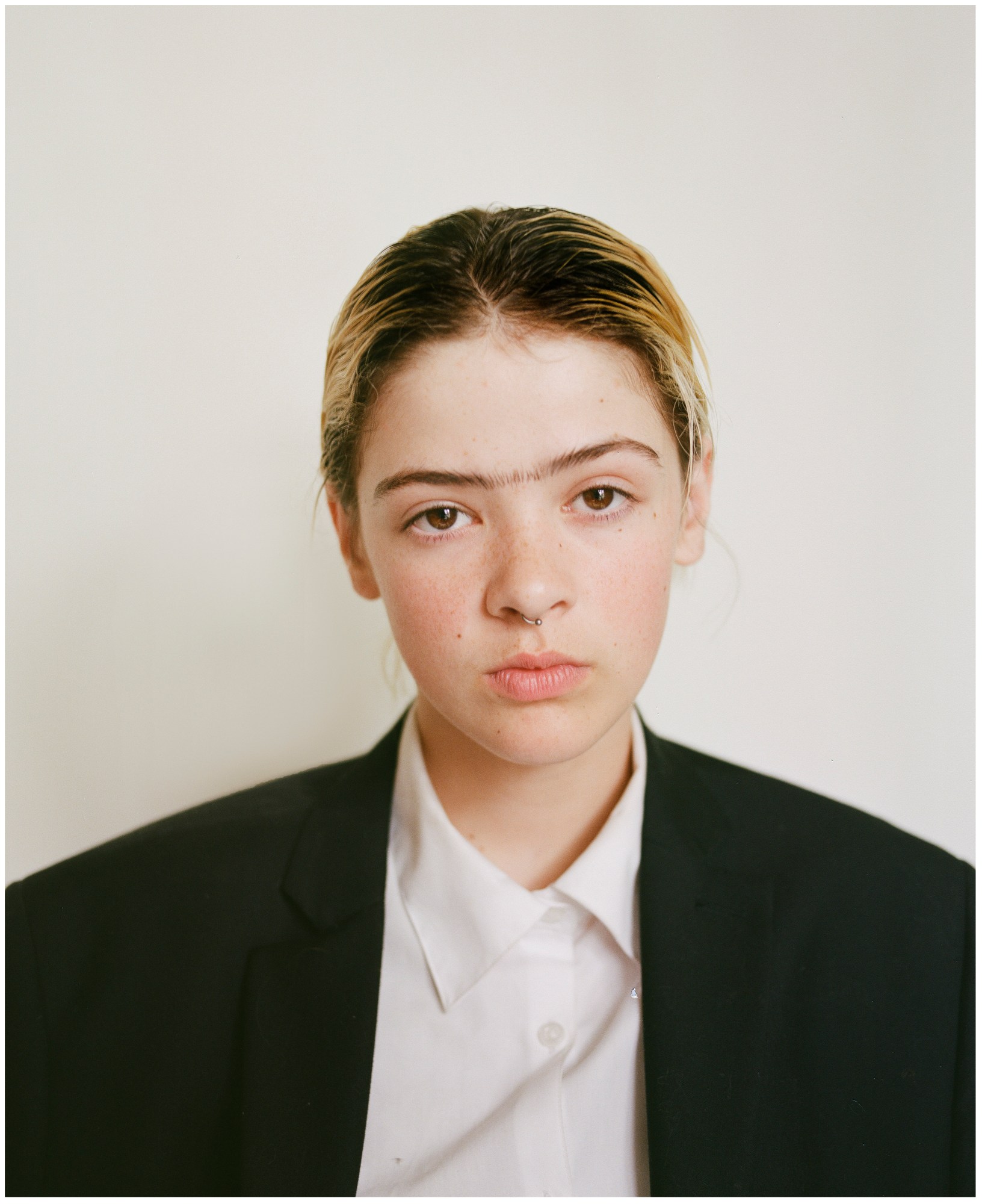 a young person with bleach blonde hair looks down the camera lens, they have a drawn-on monobrow and are wearing an oversized suit