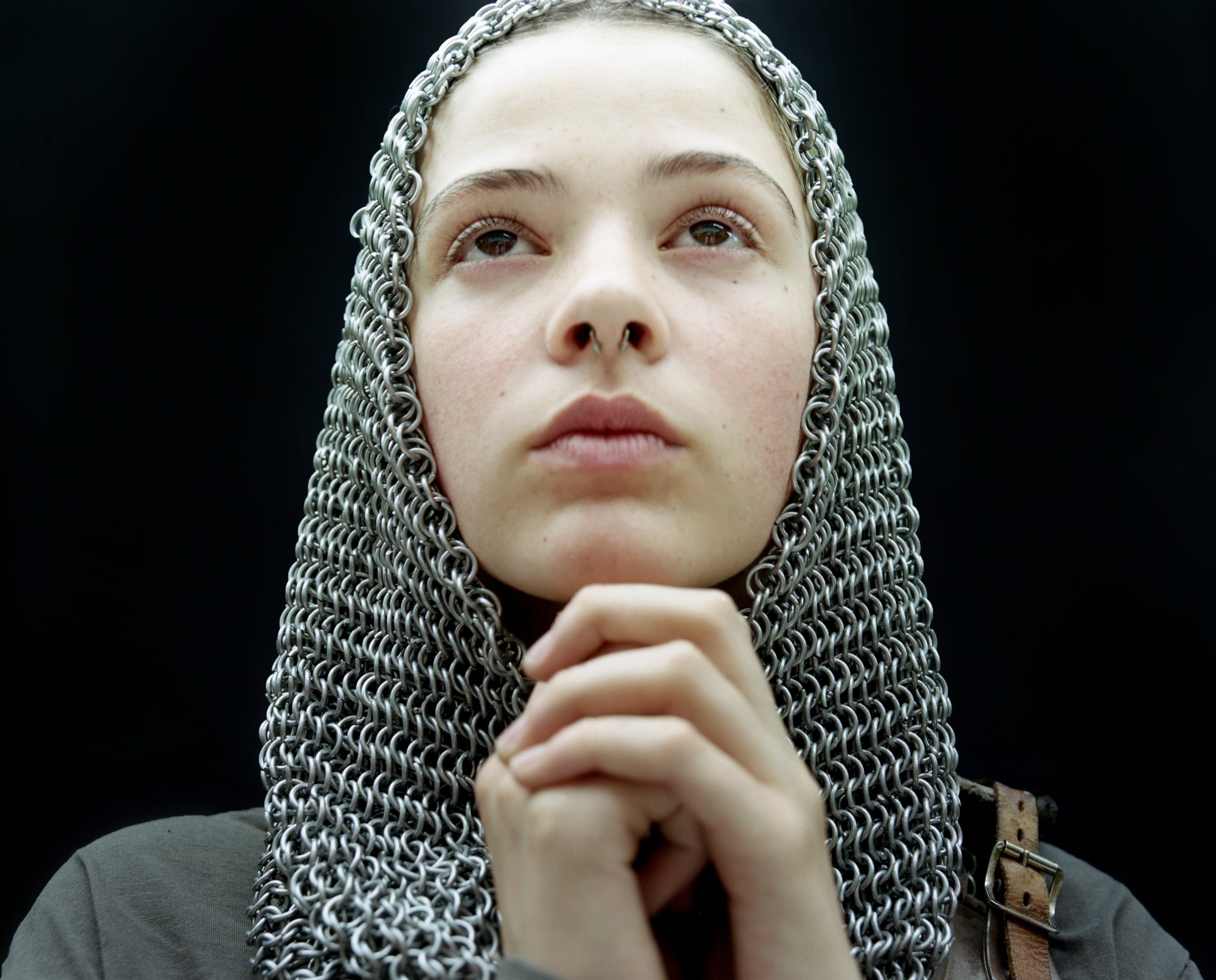 a young person with a septum piercing wears a chainmail hood; they hold their hands in prayer, joan of arc like