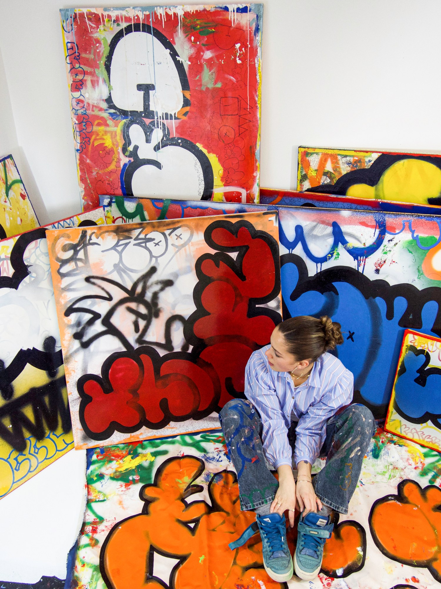 the artist franka mangano in her studio wearing pain splattered denim and her hair scraped back. she is surrounded by her paintings which are like grafitti.