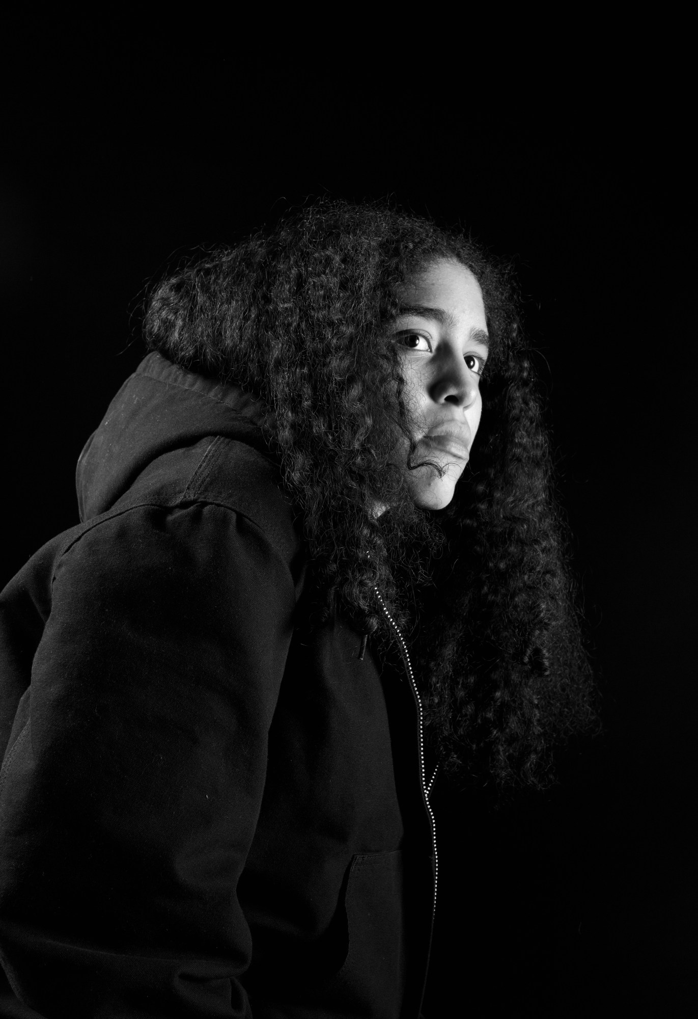 a black and white portrait of artist stephanie rodriguez. they have a black jacket and are looking side on. their hair is curly and past their shoulders.