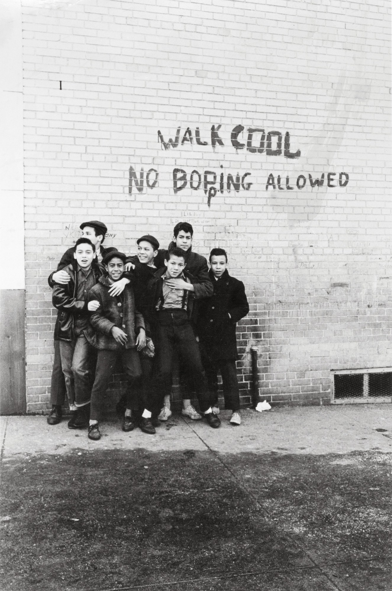 Black-and-white photograph by Steve Schapiro from the early 1960s of a group of boys holding each other and standing by a wall that says walk cool. no bopping allowed.