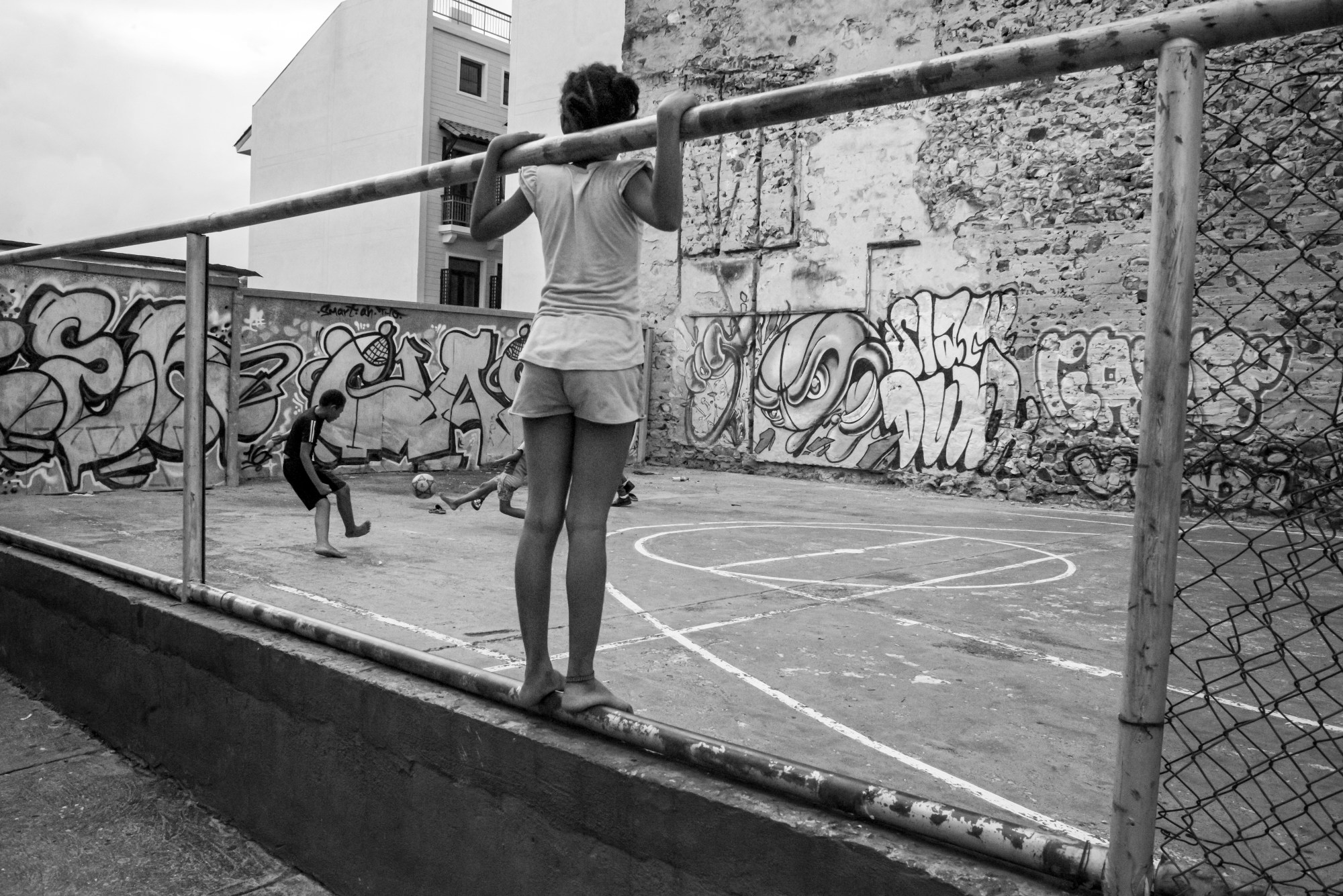 Black-and-white photograph by Theophilus Donoghue from 2019 of a girl holding onto the bars of a wire fence in a basketball playground covered in Graffiti .