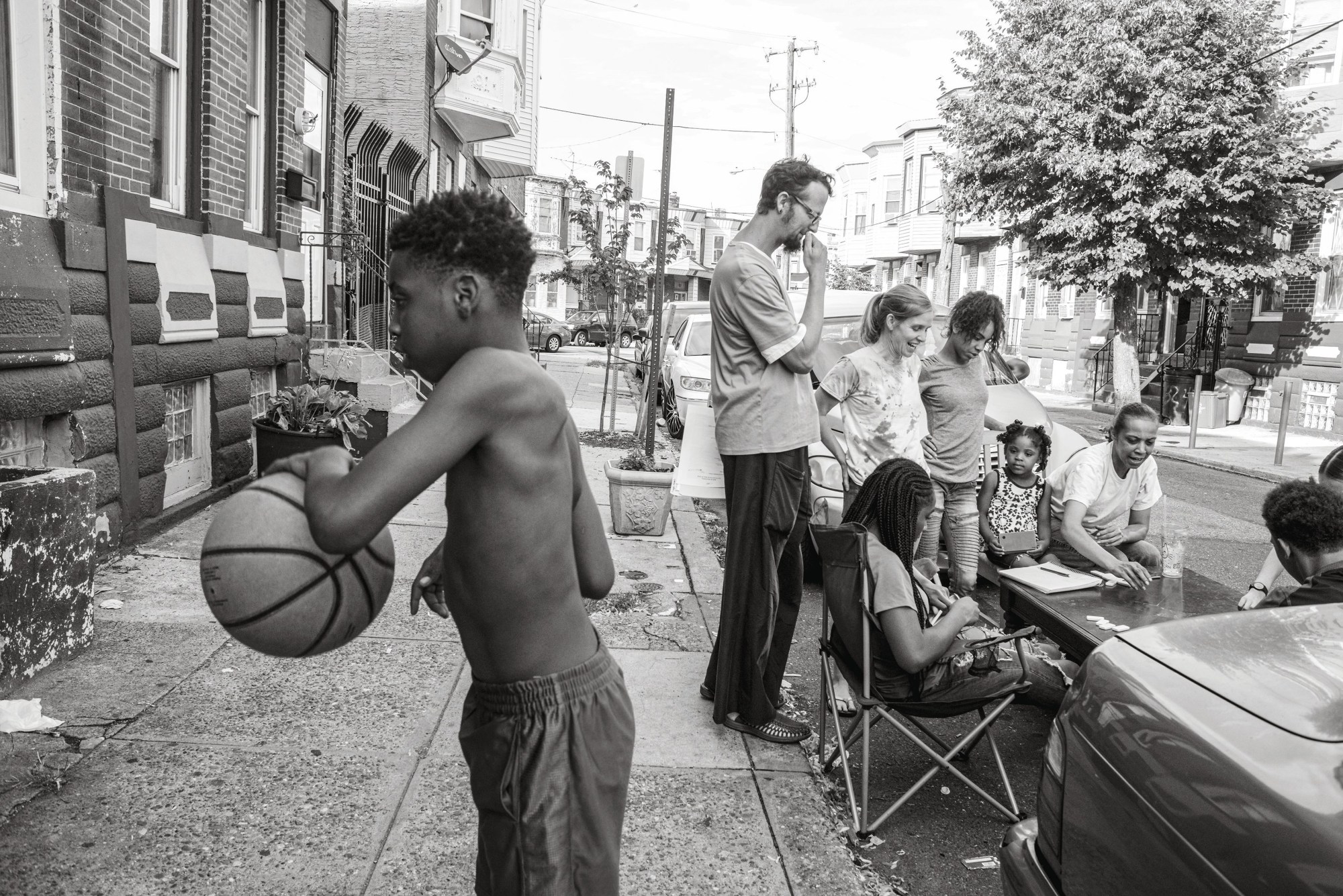 Black-and-white photograph by Theophilus Donoghue from 2018 of a boy playing basketball on a street with a family sat around on deck chairs on the road.