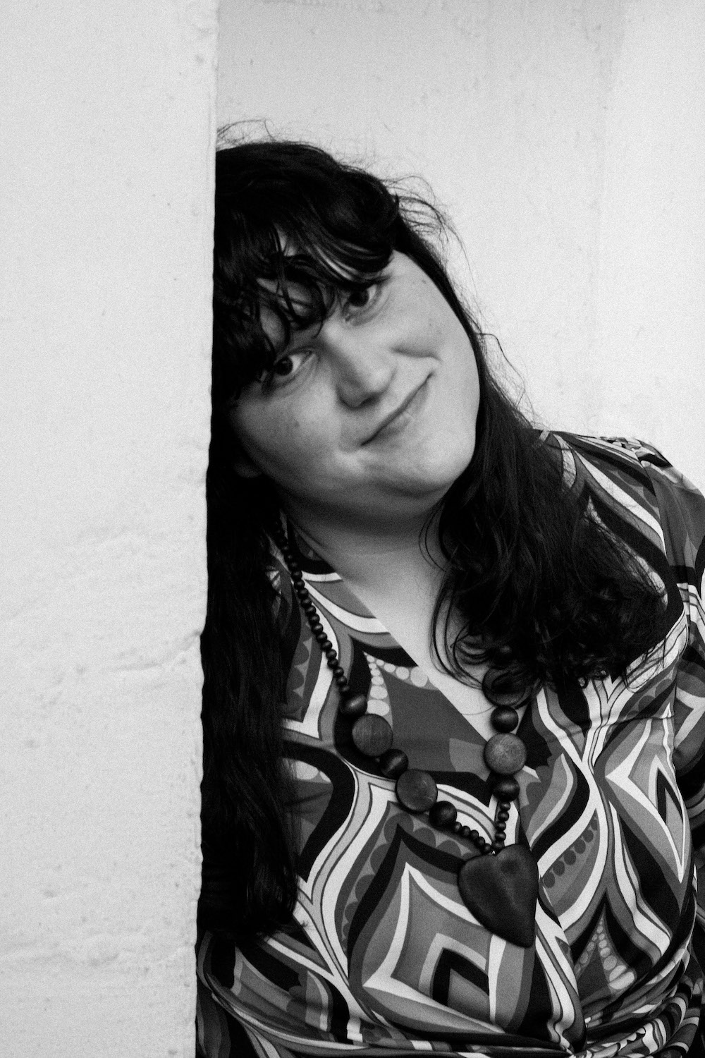 a black-and-white portrait of the author soula emmanuel, a woman with dark hair and a fringe wearing a patterned blouse and a beaded necklace. she is leaning on a wall.