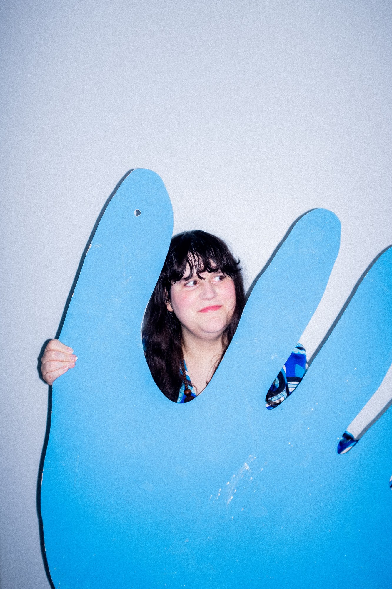 a portrait of the author soula emmanuel wearing a blue patterned shirt. she is hidden behind a massive blue cartoon hand and smiles off camera.