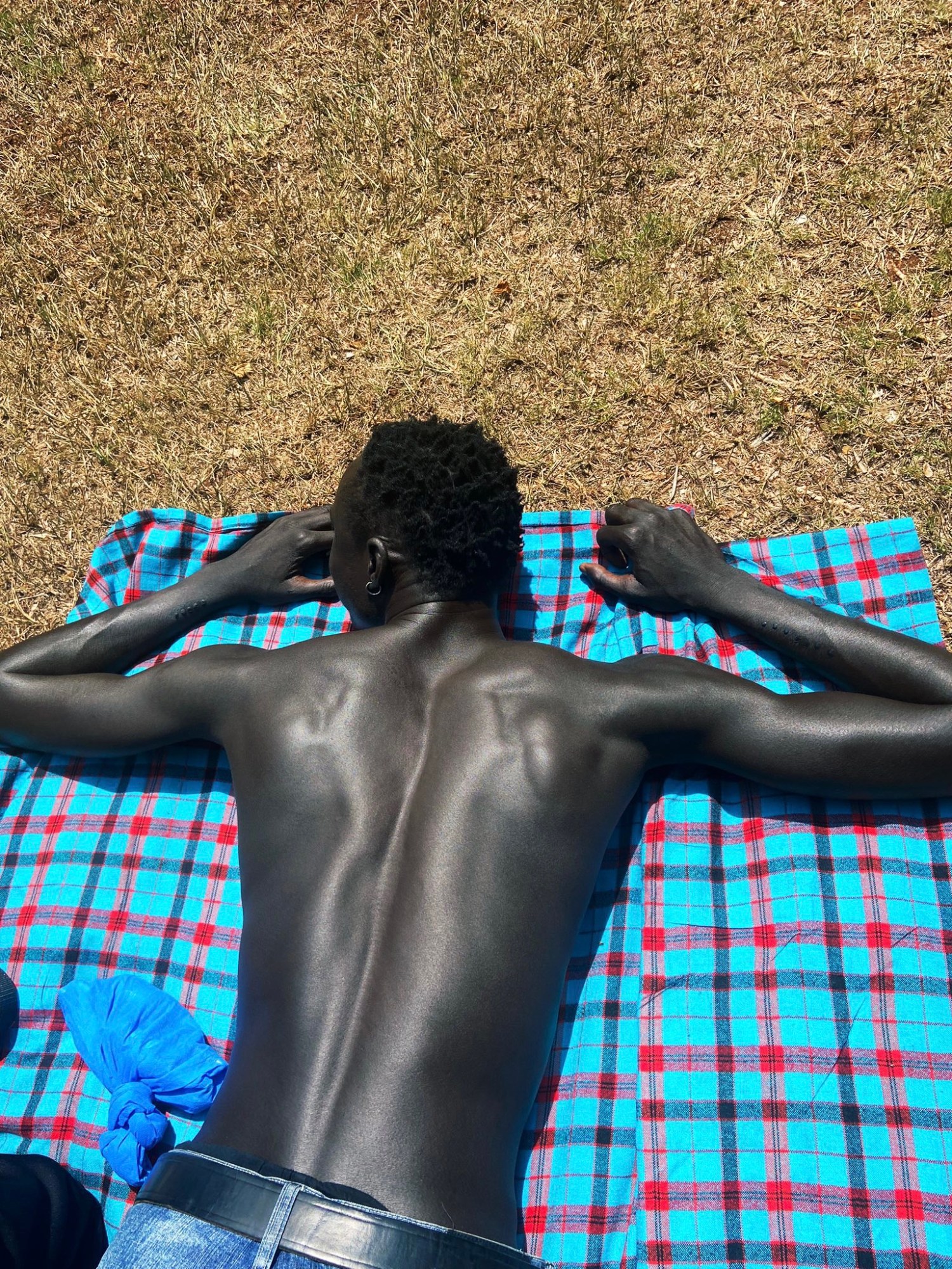 a topless man lying face down on a blue plaid picnic blanket in a field