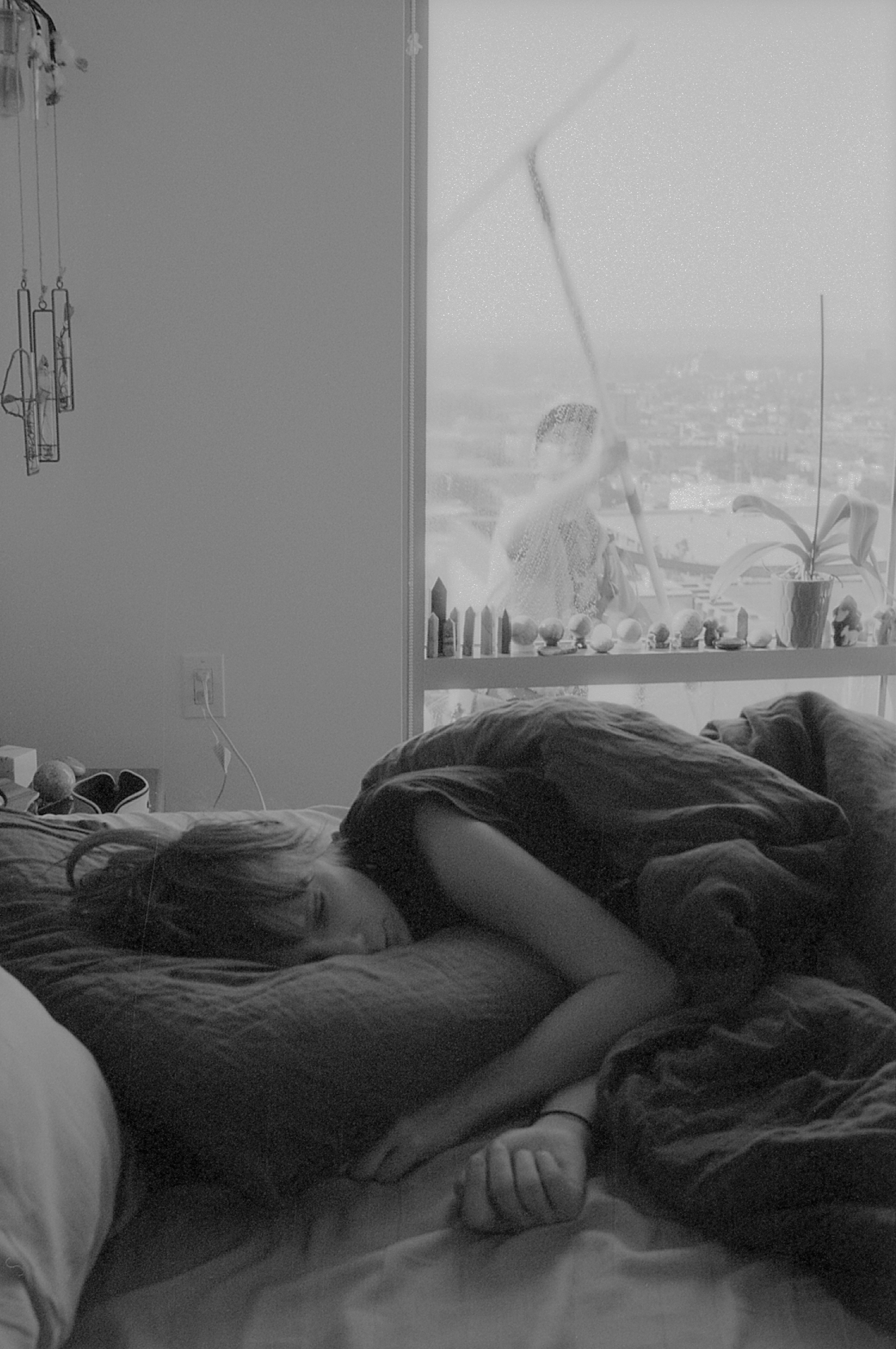 Black-and-white image of a person sleeping on their side in bed while someone washes the windows behind them