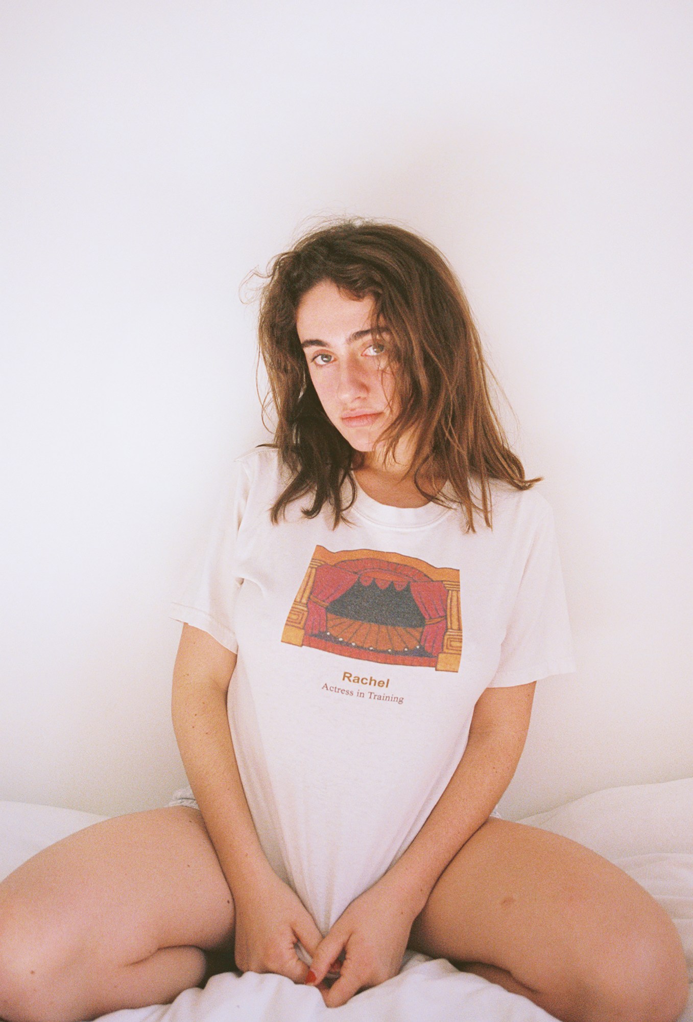 Rachel Sennott photographed by Morgan Maher sitting on a bed pulling her tshirt down between her legs.