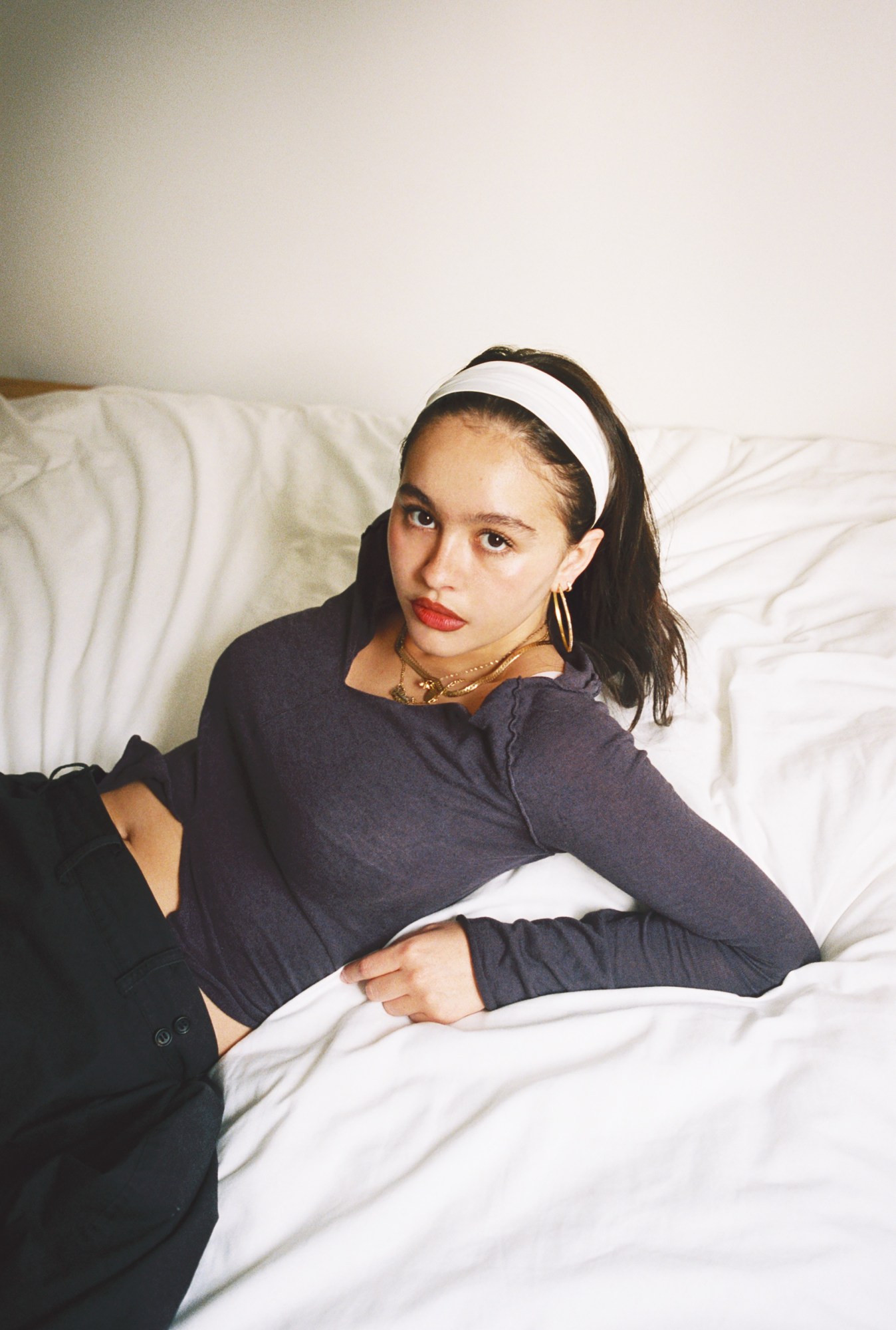 Enya Umanzor in a headband and gold jewelry photographed by morgan maher lying on a white bed