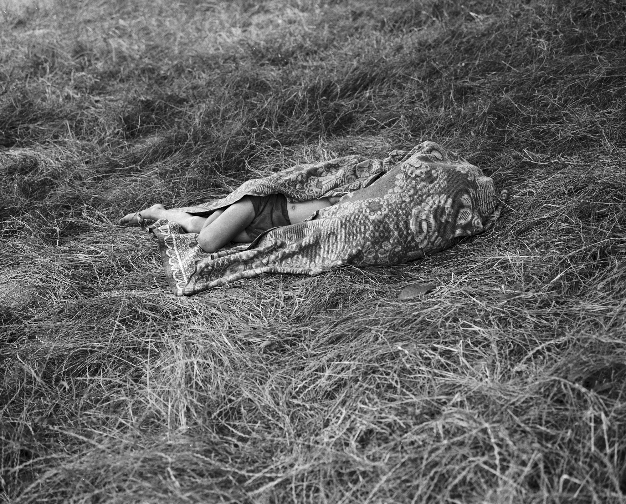 Black-and-white image of a person wrapped up in a paisley blanket lying in a field