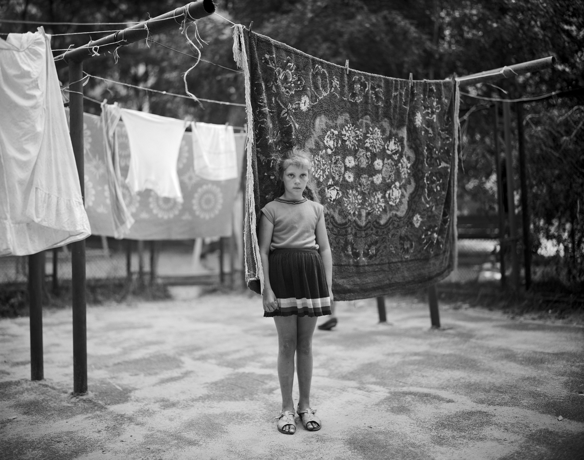 Black-and-white image of a young girl in pleated skirt and tank top standing in front of a washing line