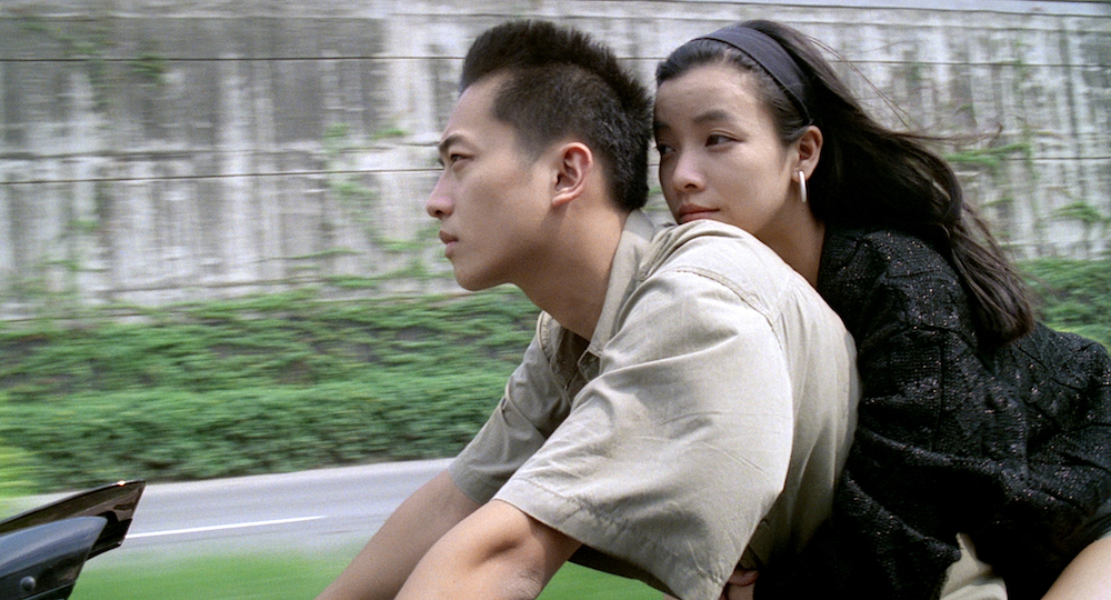 A man and a woman speeding by on a motorcycle with greenery in the background