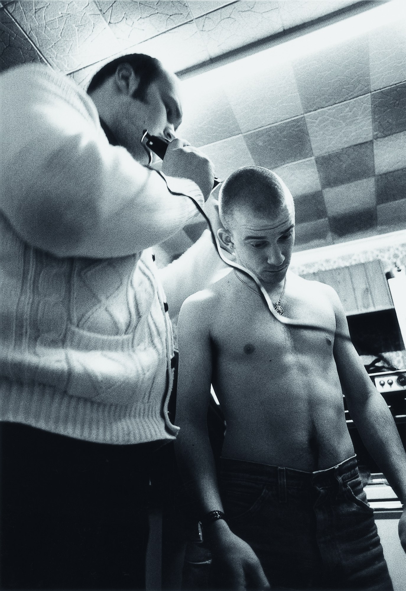 a young man has his head shaved by a bigger man in a cardigan