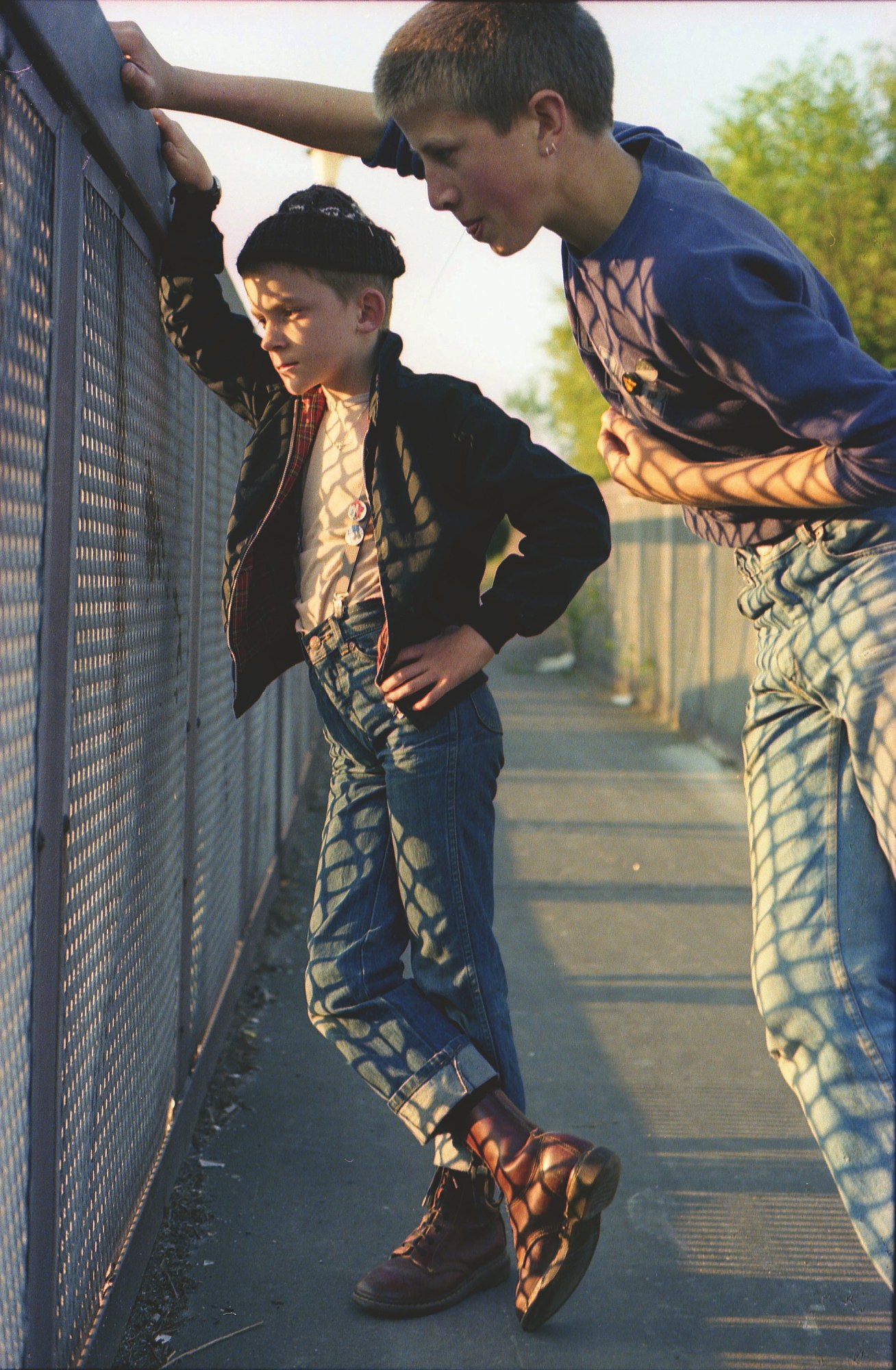 two young skinheads wearing jeans and docs stand on a bridge during golden hour