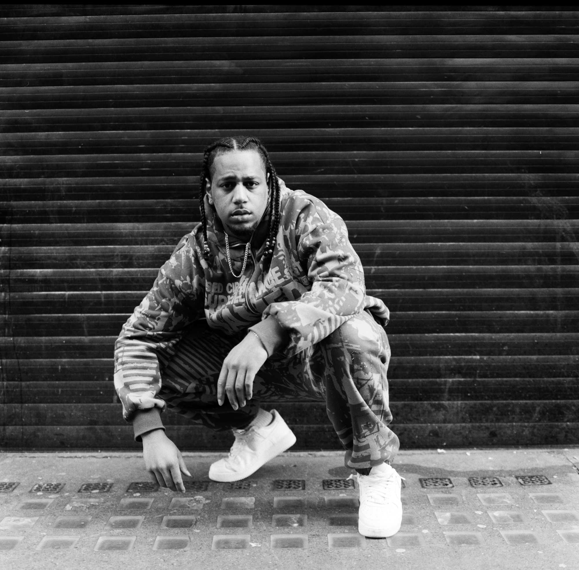 Kaine Lawrence, who plays Midnight in For Black Boys crouching in front of a grated garage door. he wears white sneakers, and a patterned hoodie sweatpant two-piece