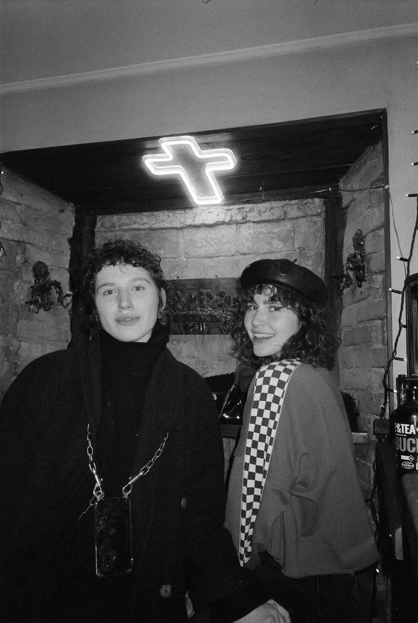 a black and white portrait of two girls with a neon cross lit up on the ceiling