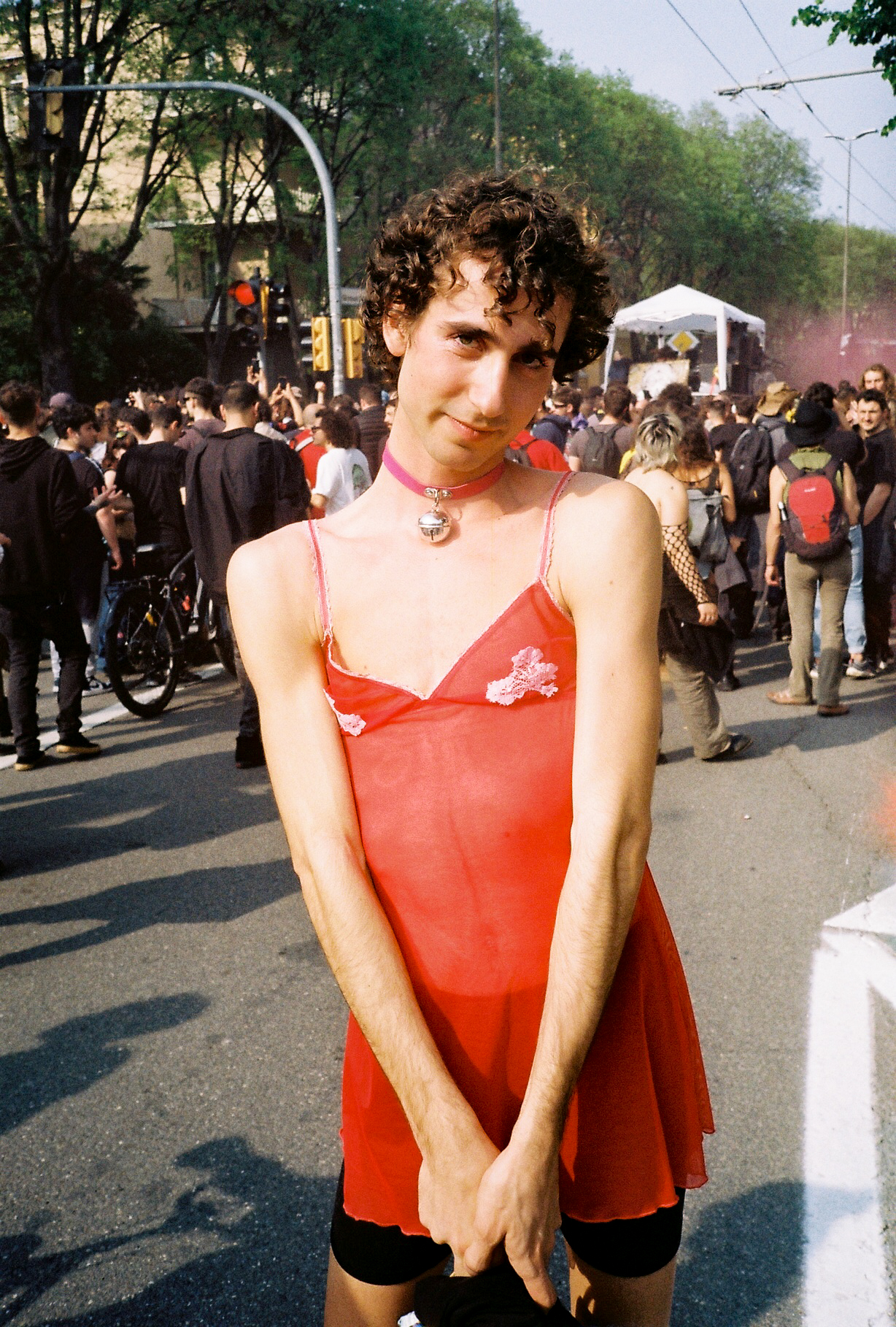 a person stands on the road with their hands clasped to their front. they're wearing bicycle shorts and a red nightgown. their hair is curly.