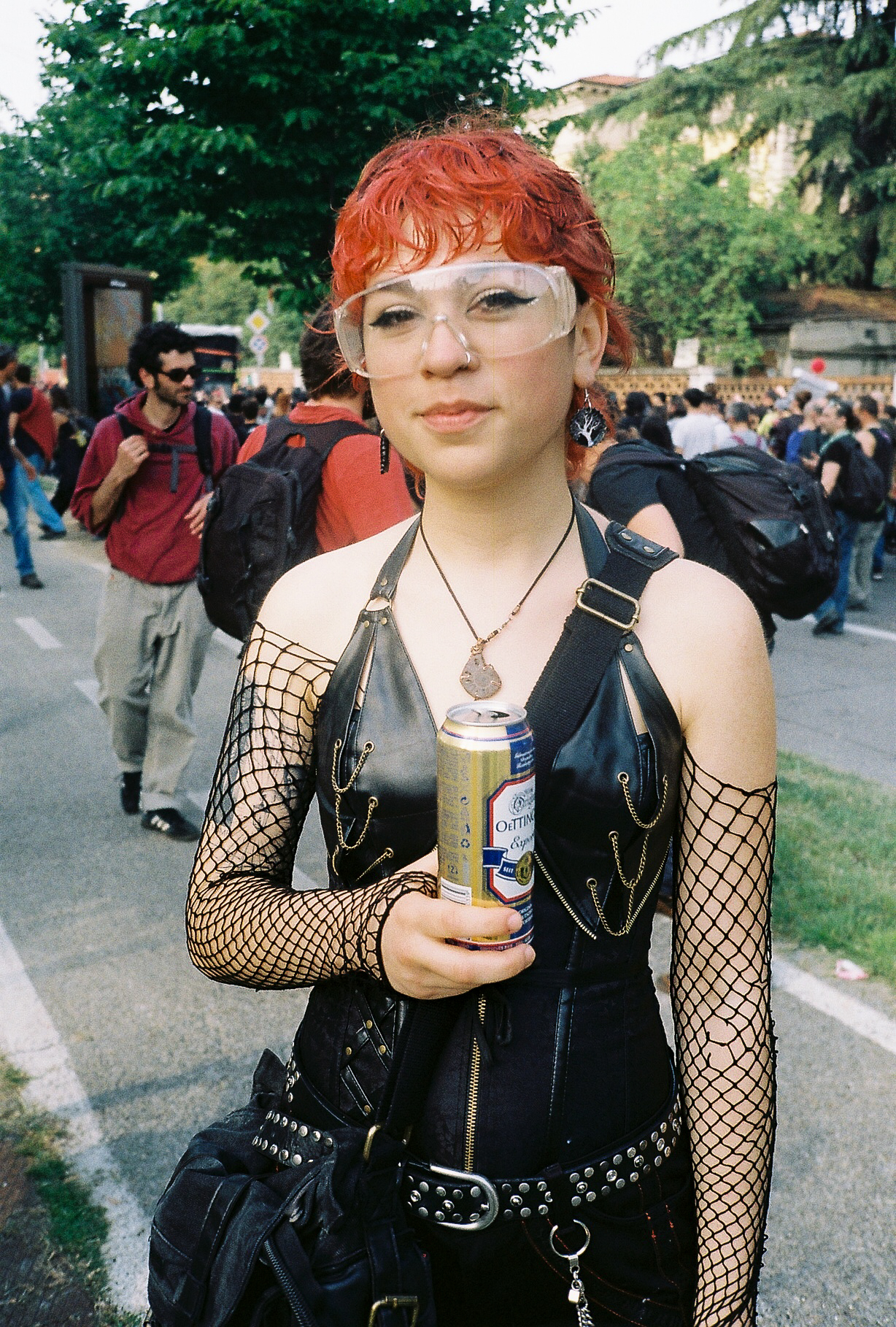 a person at a rave carries a can of beer. they are wearing a leather onepiece with a golden zip, fishnets over their arms and protective glasses. their hair is cropped and red.