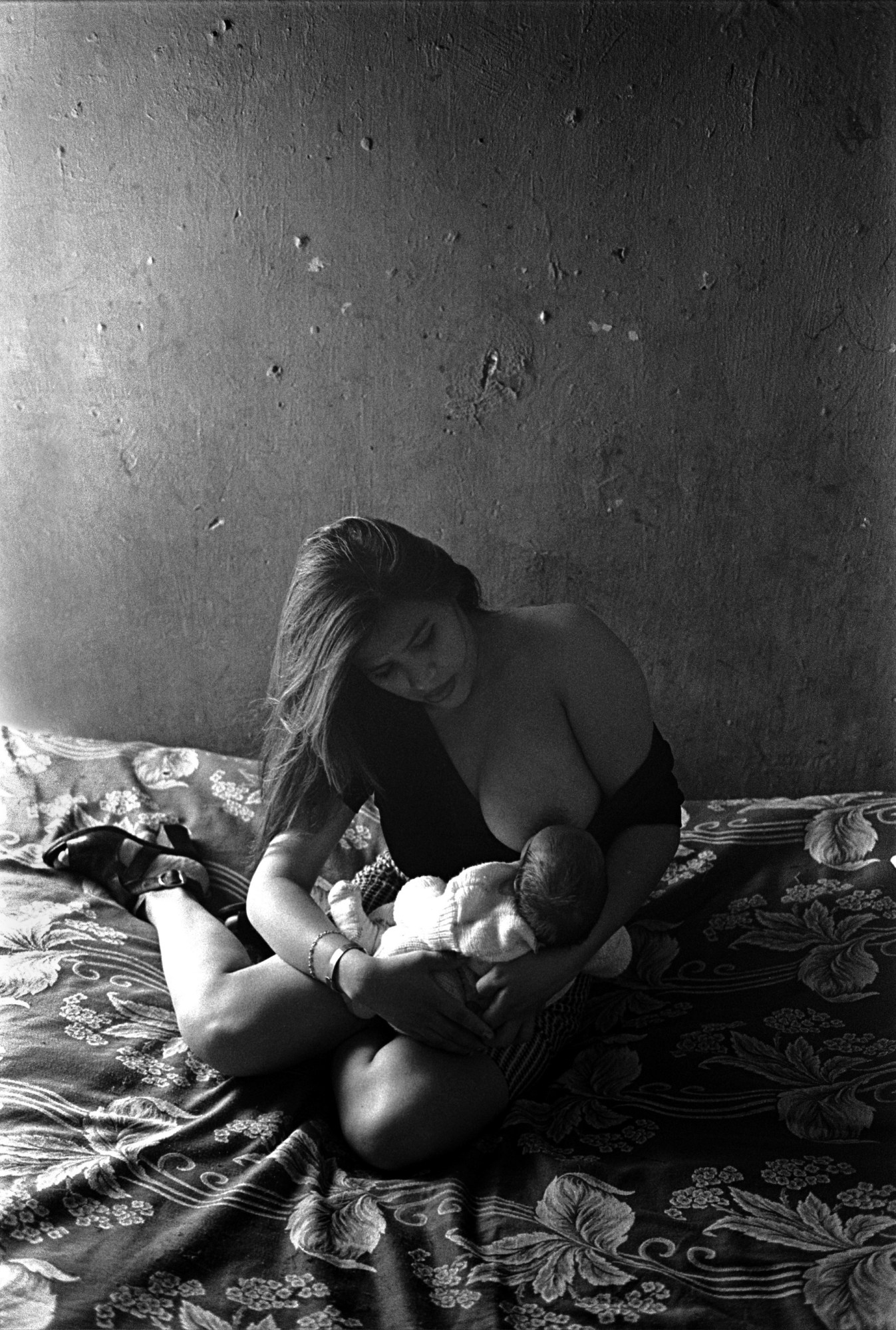 Black-and-white photograph by Joseph Rodriguez in Mexico City in the 90s of a woman breastfeeding a baby while sat on the bed.