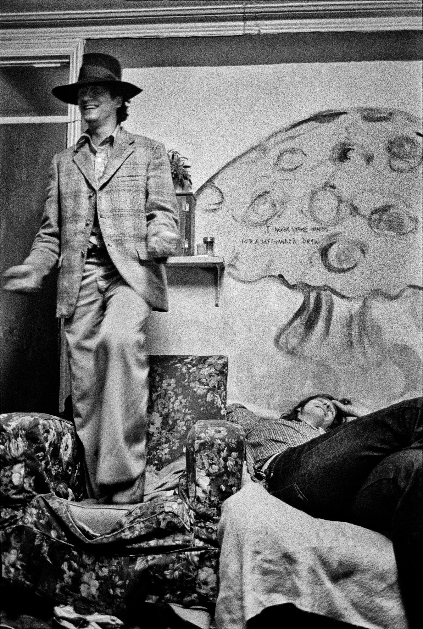 two models are laughing, one standing on an armchair and the other reclining on a bed under a large drawing of a mushroom