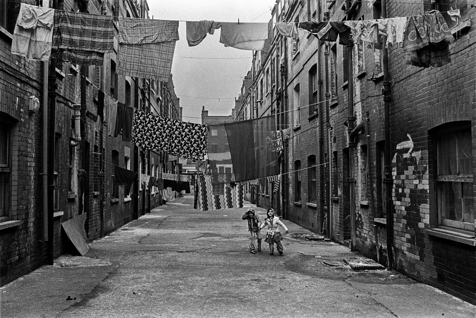 two children play outside between buildings, standing under lines of hanging laundry