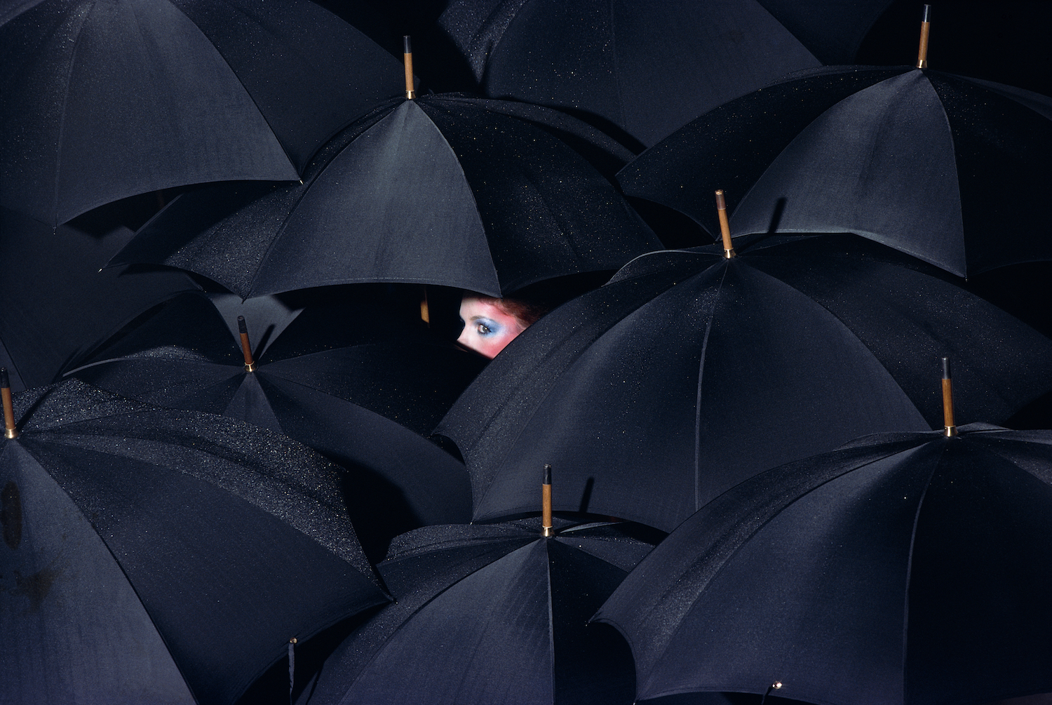 A woman in striking make-up caught under a crowd of black umbrellas