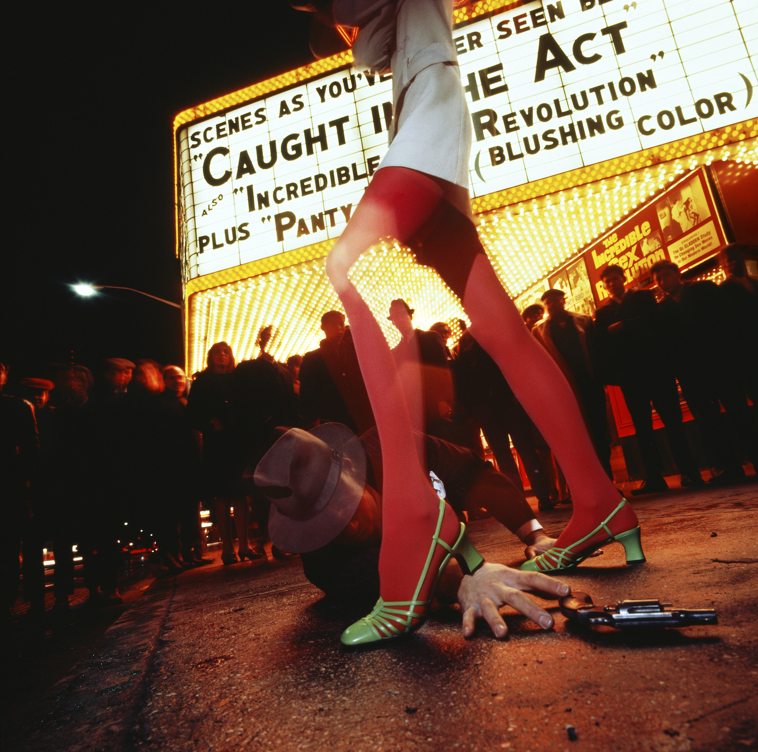 A woman in red tights and green shoes stands on a man holding a gun in front of a theatre