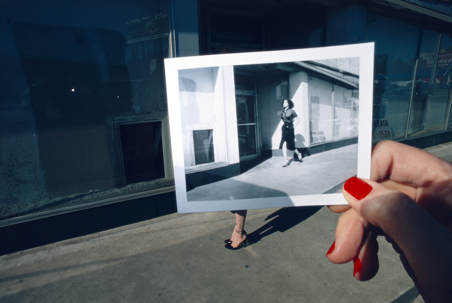 A hand with red nails holds a polaroid against a street scene