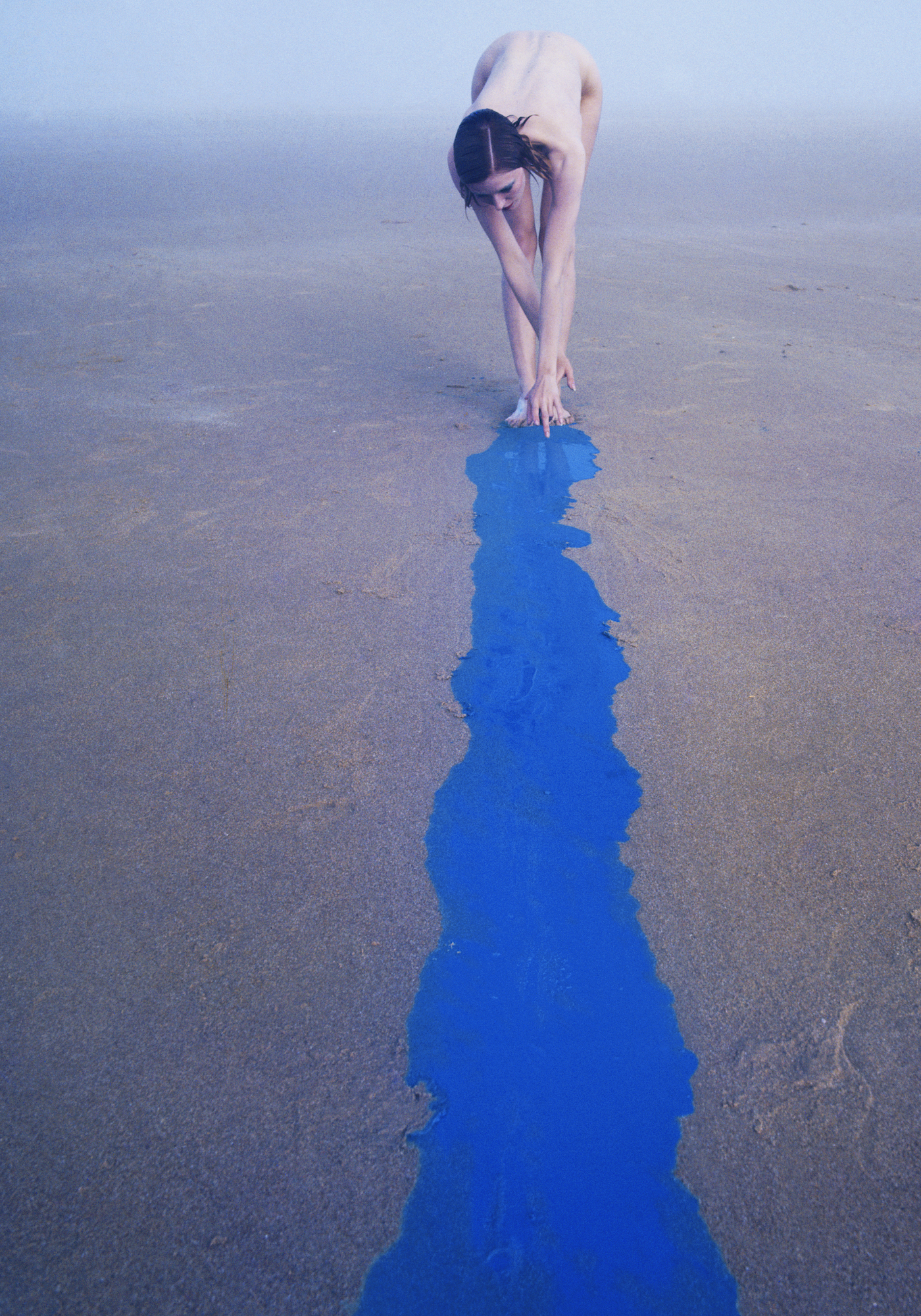 A naked woman on a beach next to a blue slick of oil