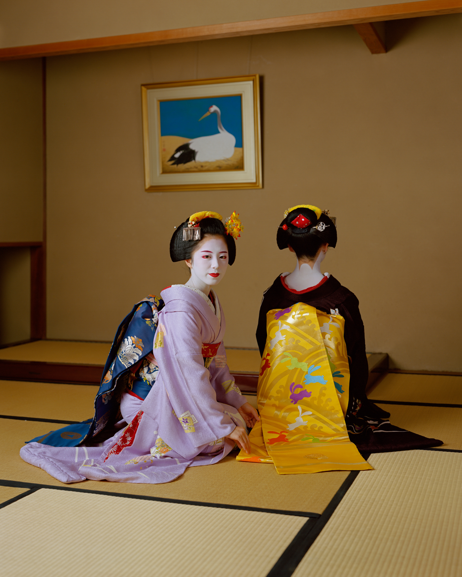 Two traditional geishas in purple and yellow kimonos sit on the floor, one of them looking back at the camera