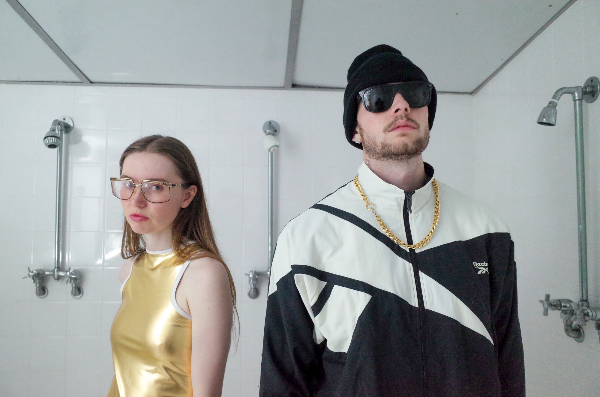 a white woman and man wearing outfits with accents of gold lame stand in a white tiled public shower room; the man is wearing dark glasses