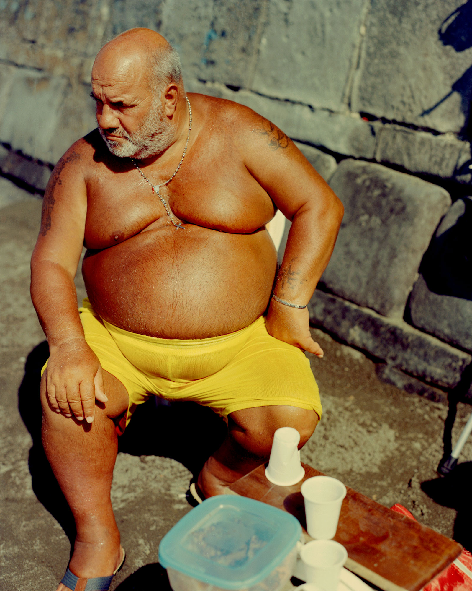 A bronze-skinned man with a bald head and beard sits shirtless on a chair on the rocky beach in Santa Lucia Naples. He's wearing yellow swimshorts and sandals, and is looking off camera