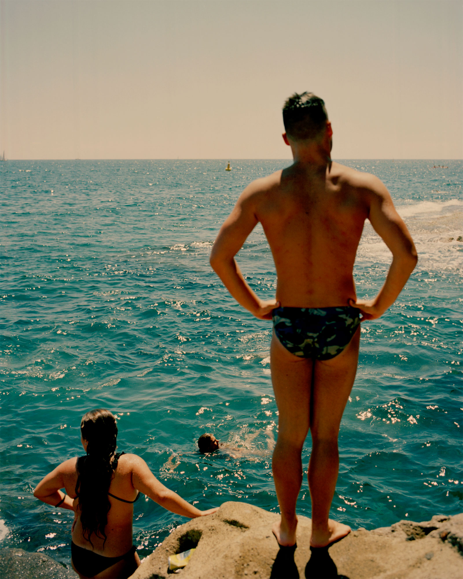 a man in swim shorts and a girl in a bikini look out onto the blue water standing on a rock. someone swims close to them.