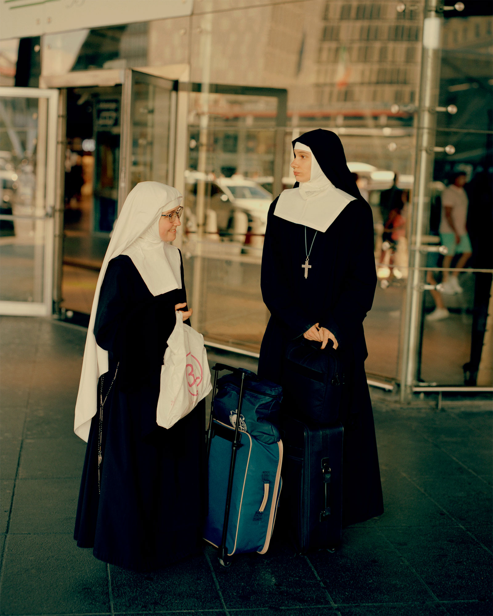 two nuns in full regalia stand on the pavement in the shade carrying blue suitcases