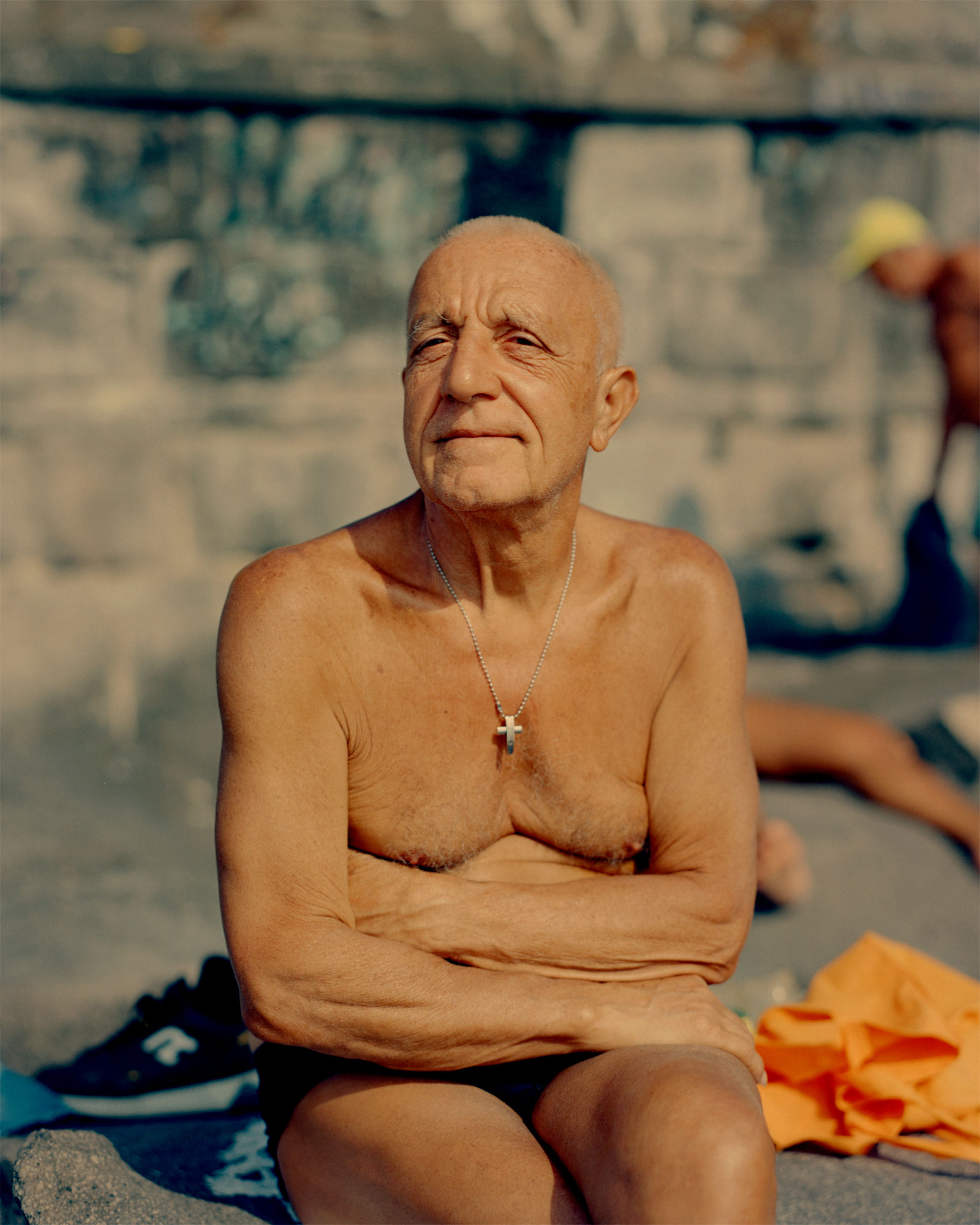 an elderly man with sagging skin sits on the beach topless wearing a cross round his neck. his skin is bronze .