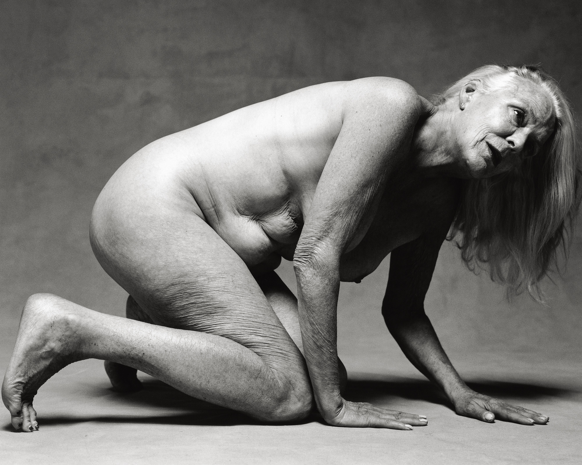 an elderly woman naked on her hands and knees. she has long grey hair over her shoulder.