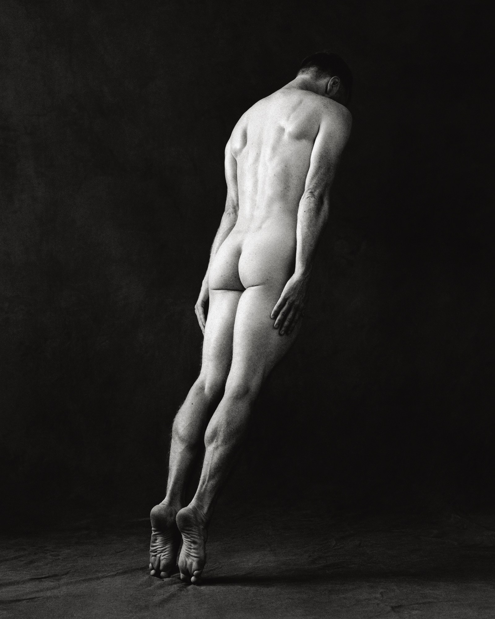 a man on his tiptoes leaning forward naked in a photo studio