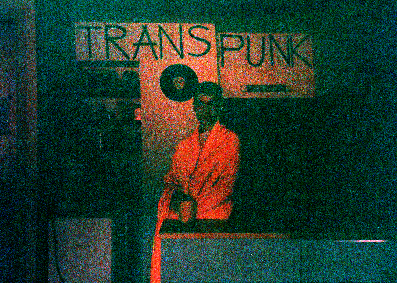 a blurry photo of a young man wrapped in a blanket standing in front of a trans punk sign