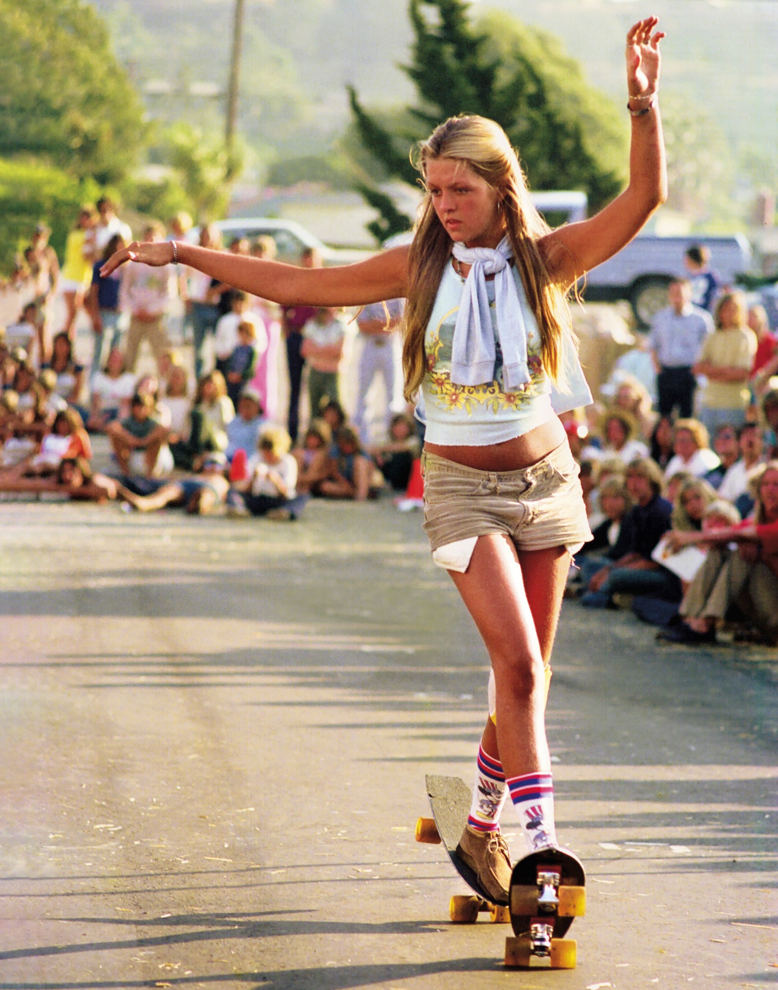 a girl in short shorts and tube socks riding two skateboards in the 70s by hugh holland