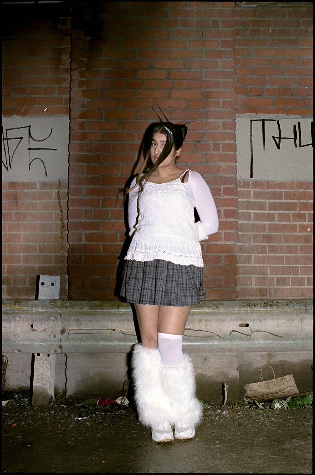 a raver in white furry socks and a pleated skirt standing in front of a wall outside