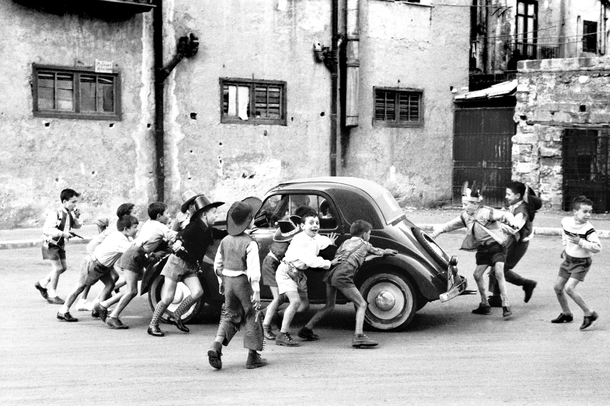 black-and-white photograph by Enzo Sellerio of kids dressed as cowboys pushing a car on the road