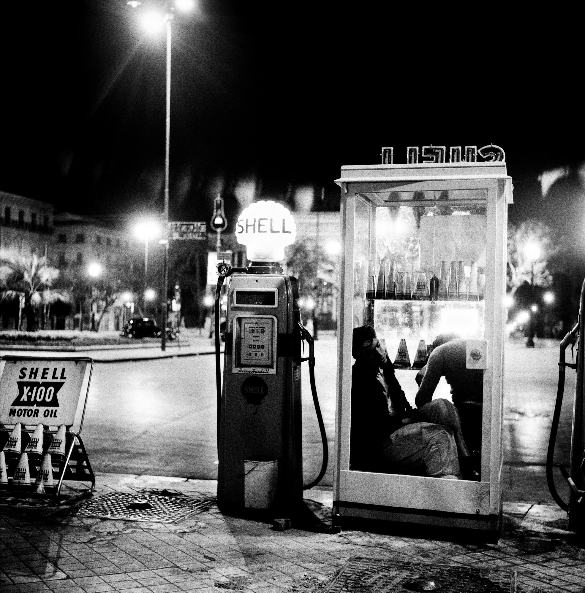 black-and-white photograph by Enzo Sellerio of a man sitting in a petrol station booth on the street.