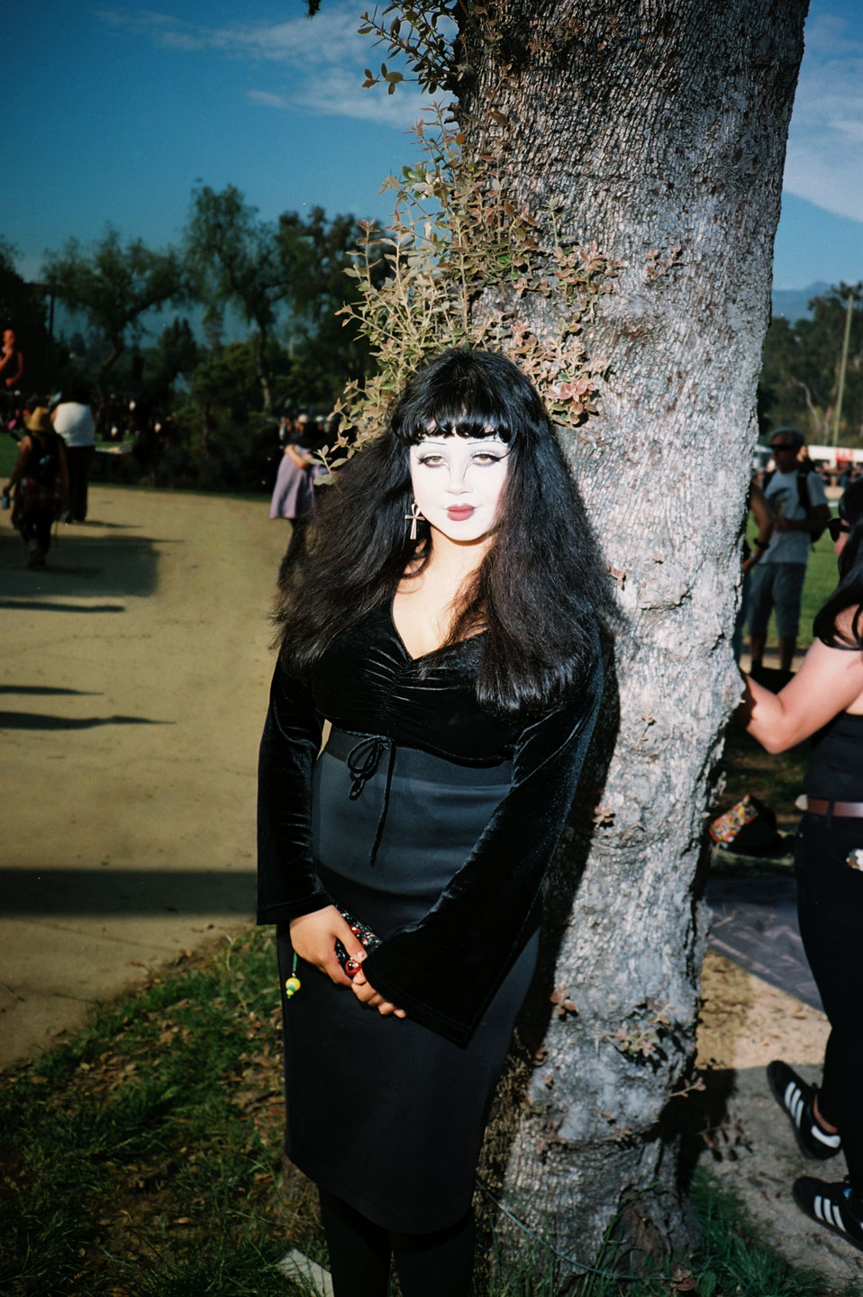 a white-faced goth girl wearing black velvet stands in front of a tree on a sunny day