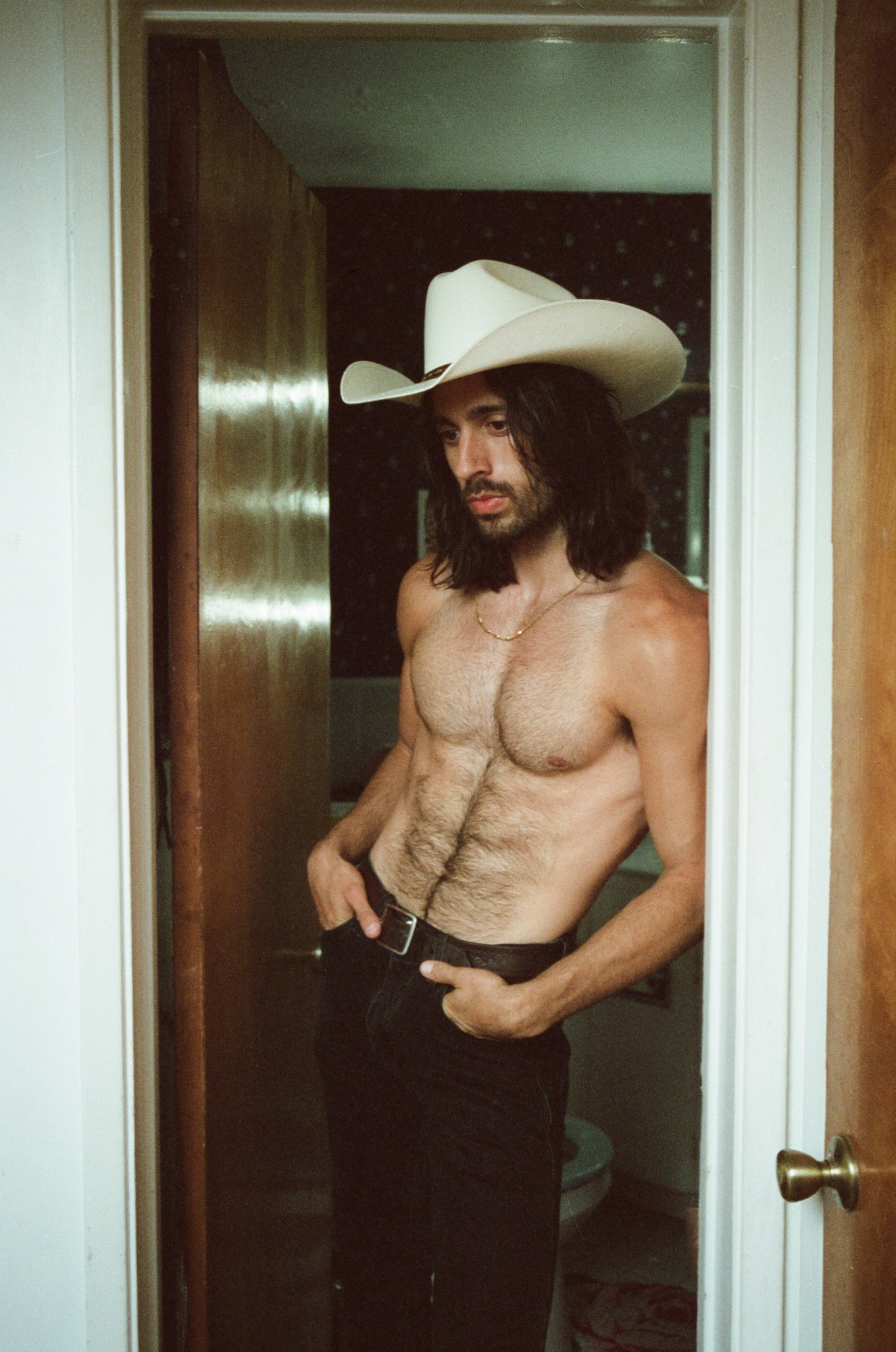 a topless man in a cowboy hat leans against a doorframe